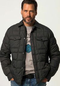 Zwarte gewatteerde jas met knoopsluiting aan de voorkant, twee borstzakken en een kraag, gedragen over een lichtgrijs T-shirt.