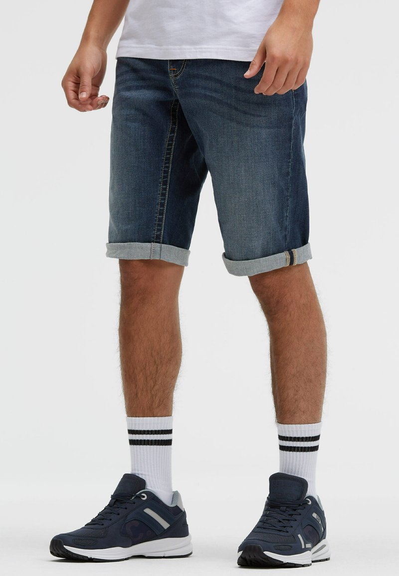 Man wearing rolled-up dark denim shorts, white socks with black stripes, and navy blue sneakers, standing on a plain white background.