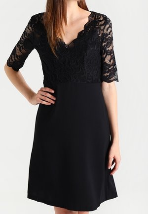 Femme portant une robe noire avec des manches courtes en dentelle et un décolleté en V festonné, debout avec une main sur la hanche.