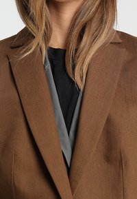Blazer marrone con un tessuto texturizzato, revers a punta e fodera grigia a contrasto. Presenta una vestibilità su misura e una silhouette pulita.
