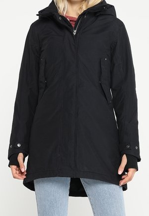 Frau trägt eine schwarze Winterparka mit Kapuze, Reißverschluss und Druckknopftaschen, kombiniert mit hellblauen Jeans.