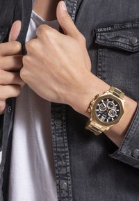 Orologio d'oro con quadrante nero, accenti argento e funzioni cronografo, indossato su un polso. Sullo sfondo si vede una camicia in denim scuro.