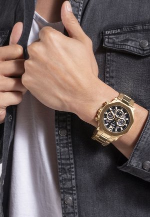 Gouden horloge met een zwart wijzerplaat, zilveren accenten en chronograaf functies, gedragen om een pols. De achtergrond bevat een donkerdenim shirt.