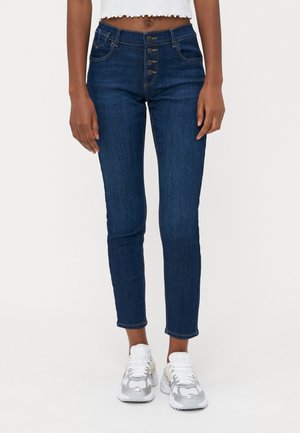 Femme portant un jean skinny taille haute bleu foncé avec quatre boutons à l’avant et des baskets blanches, sur un fond clair uni.