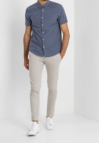 Camicia a maniche corte con bottoni in tessuto blu scuro, indossata con pantaloni slim-fit beige chiaro e sneakers bianche. Tessuto liscio e vestibilità sartoriale.