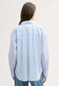 Camisa de corte amplio en algodón de color azul claro, con pliegue en la parte trasera, mangas largas, puños con botones y cuello clásico. Textura suave y ajuste relajado.