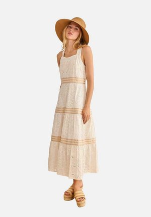 Mujer con un vestido maxi blanco de encaje sin mangas, sombrero de ala ancha color beige y sandalias de plataforma, de pie frente a un fondo liso.