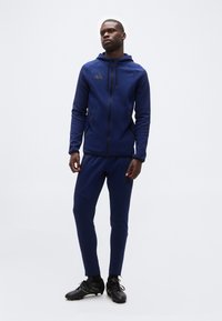 Άντρας που στέκεται φορώντας μπλε ναυτικό φούτερ με φερμουάρ, ταιριαστά παντελόνια φόρμας, μαύρα παπούτσια ποδοσφαίρου με πράσινες λεπτομέρειες σε λευκό φόντο.