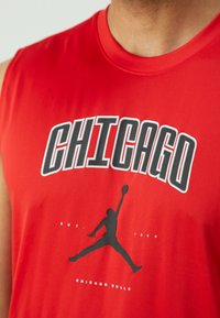 Piros ujjatlan sportpóló, amelyen a "CHICAGO" felirat fehér és fekete színben szerepel, valamint egy fekete sziluett grafika egy kosárlabdázóról.