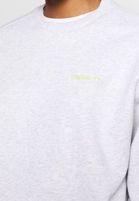 Šedá mikina s kulatým výstřihem z měkké látky, s malým neonově žlutým logem Carhartt na hrudi a klasickým žebrovaným límcem.