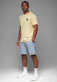 Lys gul bomulds T-shirt med et rundt logo på brystet, parret med lyseblå shorts og hvide sneakers med sorte accenter.