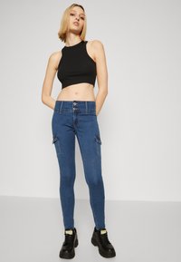 Pantalones cargo ajustados de mezclilla azul con dos bolsillos laterales, una cintura alta y un cierre de doble botón. Combinados con un top corto sin mangas negro.