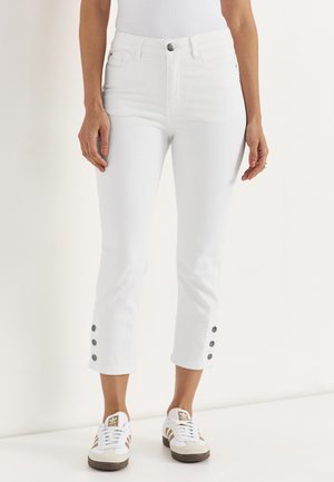 Vrouw draagt witte cropped jeans met drie zwarte knopen aan de buitenkant van de onderbenen en witte sneakers met bruine strepen.