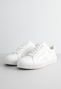 Zapatillas blancas con una parte superior de cuero suave, acentos texturizados en los laterales y una suela de goma con una textura rugosa. Cordones visibles en la parte superior.