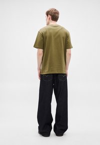 Olivno zelena bombažna majica z okroglim ovratnikom, združena z ohlapnimi temno modrimi kavbojkami iz jeansa, ki imajo vidne zadnje žepe in kontrastne šive.