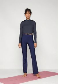 Mörkgrå tröja med långa ärmar och marinblå yogabyxor på en person som står på en ljusrosa yogamatta, med en figurnära design och slät yta.