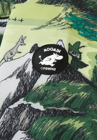 Zielony materiał z ilustracjami Muminków, z wyraźną czarną okrągłą łatką z logo Muminków i postacią na teksturowanym tle.