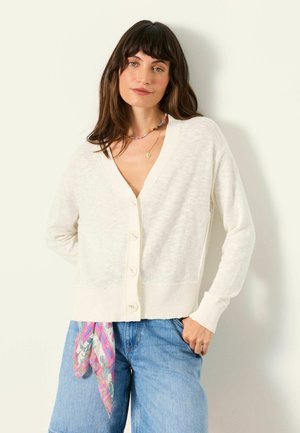 Kvinde iført en hvid knappet cardigan, løstsiddende blå jeans med et farverigt tørklæde og lagdelte halskæder, stående mod en lys baggrund.