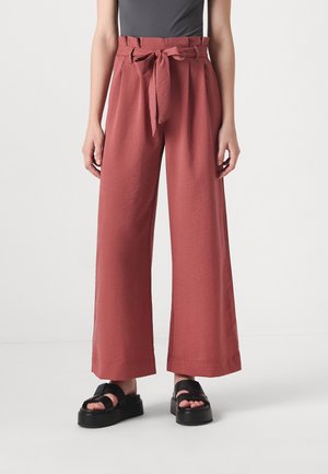 Donna che indossa pantaloni a gamba larga a vita alta di colore ruggine con cintura annodata, top nero senza maniche e sandali platform neri.