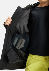 Osoba w czarnej bluzce i żółtych spodniach, trzymająca otwartą czarną kurtkę, aby pokazać wewnętrzną siatkową kieszeń zawierającą złożone papiery.