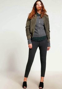 Olive green bomber jacket over a textured gray zip-up sweater, paired with black slim-fit pants and black slide sandals.