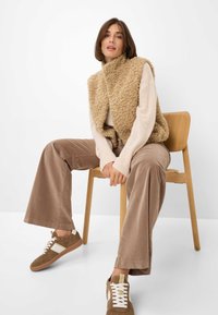 Beige fuzzy vest over een crèmekleurige trui, gecombineerd met lichtbruine ribfluwelen broek en bruine sneakers met witte accenten, zittend op een houten stoel.