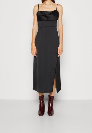 Abito midi in satin nero con spalline sottili, scollatura a cowl, dettaglio con spacco laterale e corpetto aderente, abbinato a stivaletti alla caviglia color bordeaux.