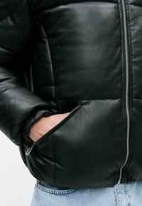 Jachetă puffer din piele neagră cu fermoar în față, manșete elastice și buzunare laterale. Prezintă o textură netedă și un model matlasat.