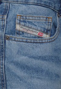 Zila džinsu bikses ar aizmugures kabatu, kurai ir balta un sarkana etiķete ar uzrakstu "Diesel Industry" un redzamām dzeltenām šuvēm.