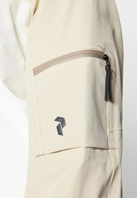 Beige jakke med en struktureret overflade, der har en lynlås-lomme og et sort logo. Designet inkluderer kontrastsyer og en slank silhuet.