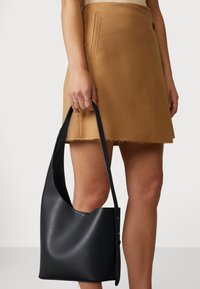 Borsa a mano in pelle nera con un design curvo e senza cuciture e tracolla lunga, sostenuta da una mano, abbinata a una gonna color cammello con orlo sfrangiato.