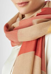 Écharpe en tissu tissé présentant une base beige avec de grands carreaux rouges et moutarde. Texture douce avec un design léger, qui se drape parfaitement.