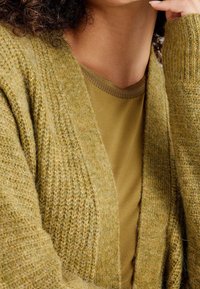 Olivově zelený kardigan z texturovaného úpletu s otevřeným předním dílem, vrstvený přes odpovídající olivově zelené tričko s žebrovaným kulatým výstřihem.