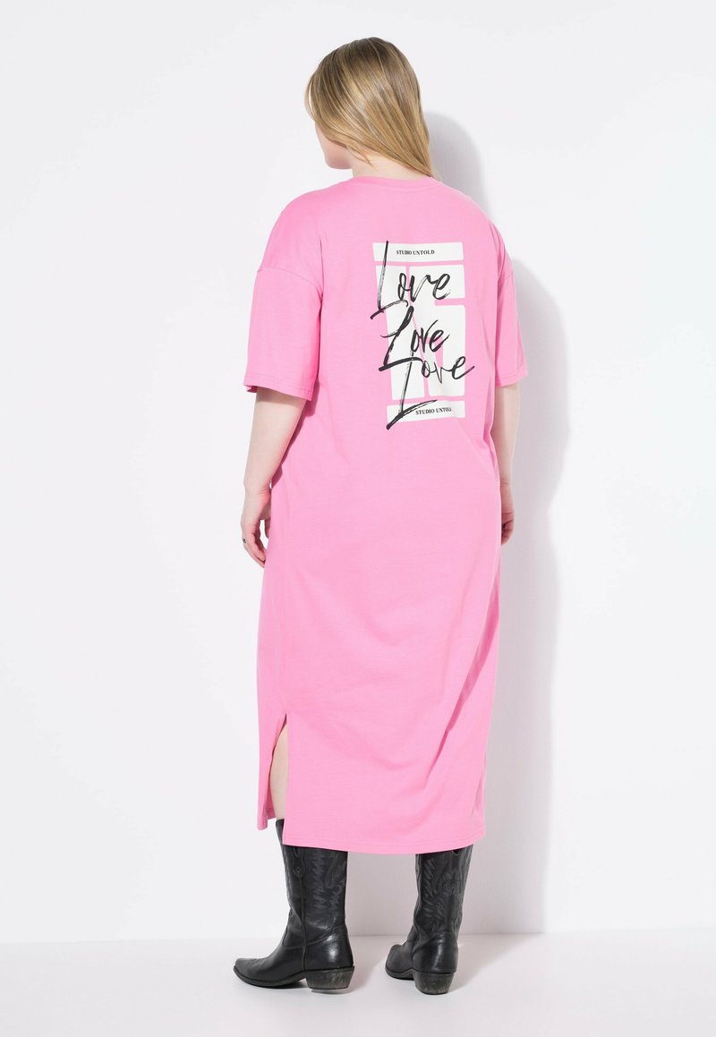 Różowa bawełniana sukienka T-shirt z krótkimi rękawami, bocznymi rozporkami i czarnym nadrukiem z napisem "Love" na plecach, zestawiona z czarnymi, wysokimi do kolan butami.