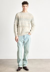 Beżowy sweter na drutach z niebiesko-brązowymi geometrycznymi wzorami, okrągły dekolt, teksturowana tkanina; w zestawieniu z jasnoniebieskimi dżinsami i ciemnymi butami.