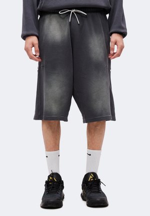 Personne portant un short long gris foncé texturé avec un cordon blanc, des chaussettes blanches montantes et des chaussures de sport noires avec des accents dorés.