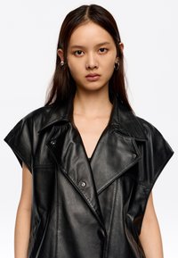 Melnas ādas veste ar pārspīlētu apkakli, īsām piedurknēm un spiedpogām. Gluda tekstūra un minimālistisks dizains.