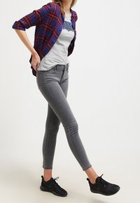 Jeans skinny grises combinados con una camisa gris que presenta un gráfico azul oscuro, superpuesta con una camisa a cuadros roja y azul marino. Zapatillas atléticas negras.