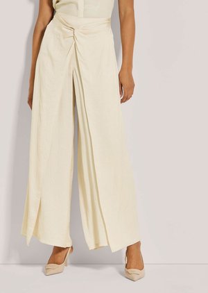 Femme portant un pantalon ample couleur crème avec un drapé sur le devant et des talons pointus beiges, debout devant un fond neutre.