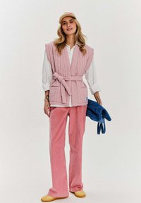 Różowa pikowana kamizelka z paskiem, biała koszula, różowe spodnie z korduroy oraz żółte buty wsuwane; trzyma niebieską torbę. Gładka struktura na całej powierzchni.