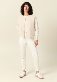 Beige fleece vest met drukknoopsluiting; gedragen over een lichte trui en witte high-waisted broek; gecombineerd met witte en tan sneakers.
