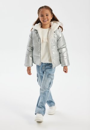 Silver puff-jacka med en pälskantad huva, kombinerad med en lätt tröja och blå cargobyxor. Modellen har på sig vita sneakers.