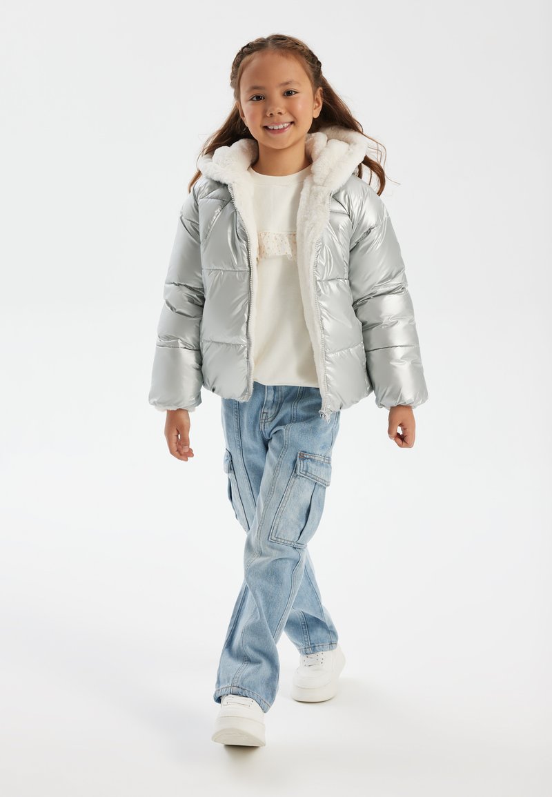 Silver puff-jacka med en pälskantad huva, kombinerad med en lätt tröja och blå cargobyxor. Modellen har på sig vita sneakers.