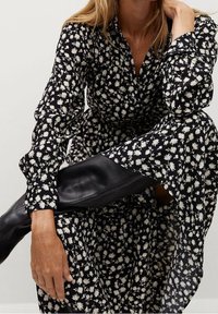 Femme portant une robe noire à imprimé floral blanc et des bottes en cuir noires jusqu'aux genoux, posant avec une jambe levée et la main reposant sur le genou.