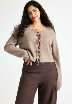 Donna con capelli lunghi e ricci indossa un maglione beige lavorato a maglia con fiocchi frontali e pantaloni marrone scuro a vita alta, posando con la mano sul fianco.