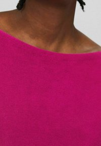 Personne portant un haut en tricot rose vif avec un col large, montrant le haut de la poitrine et une partie des cheveux tressés.