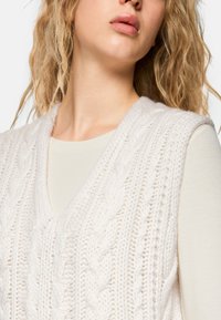 Blonde woman wearing a cream cable-knit sleeveless sweater vest over a light beige long-sleeve top, shown from shoulders to lips.