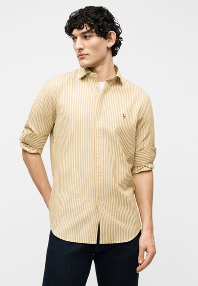 Νεαρός άνδρας με κοντά σγουρά σκουρόχρωμα μαλλιά, που φοράει μπεζ και λευκό ριγέ πουκάμισο με μακριά μανίκια που έχουν ανασηκωθεί και σκοτεινό παντελόνι.