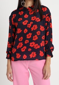 Blusa blu navy con motivo floreale rosso, maniche lunghe, scollatura rotonda e polsini arricciati, abbinata a pantaloni rosa. Texture del tessuto liscia.
