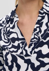 Kvinna klädd i en collared skjorta med marinblå och vit abstrakt vågig mönster och djup V-ringning, som visar övre bröstet och halsen.